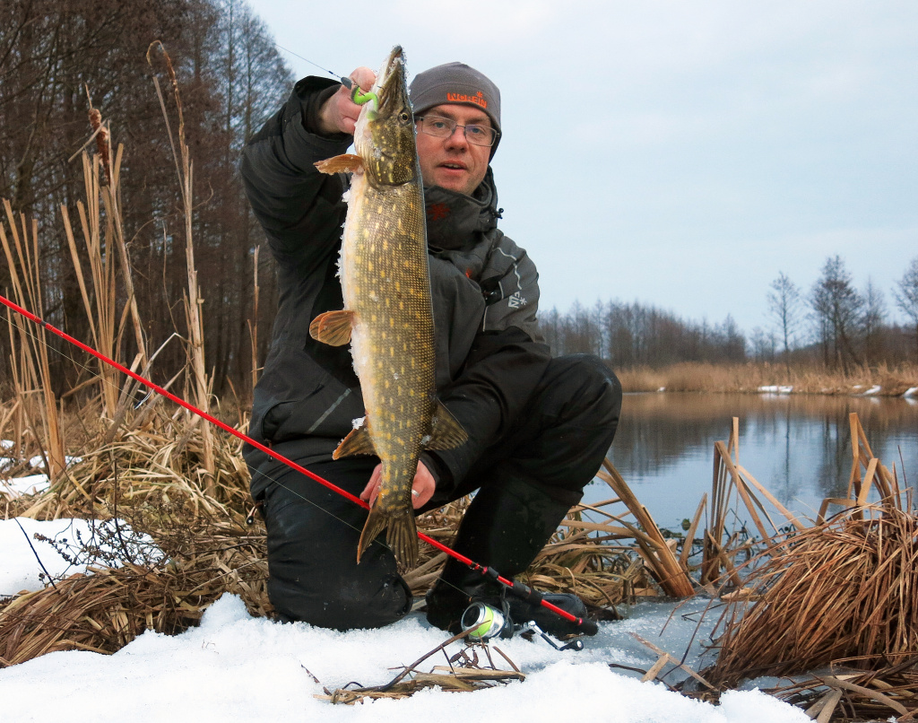Рыбак на берегу зимней реки с пойманной на спиннинг щукой