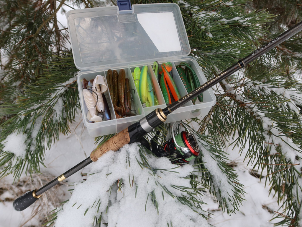 Приманки в открытой коробочке стоят на ветке заснеженной сосны, рядом стоит спиннинг